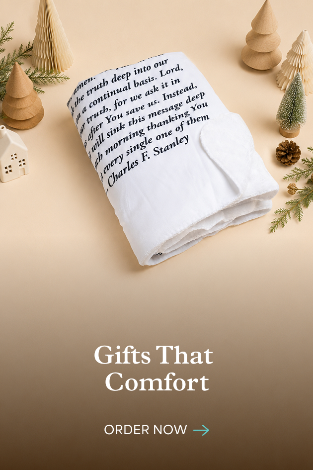 A folded white blanket printed with a Charles F. Stanley prayer, surrounded by small paper Christmas trees. Gifts that comfort. Order now.