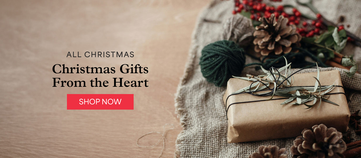 A gift wrapped in brown paper and twine, with a pinecone, red berries and green yarn on burlap. All Christmas. Christmas gifts from the heart. Shop now.